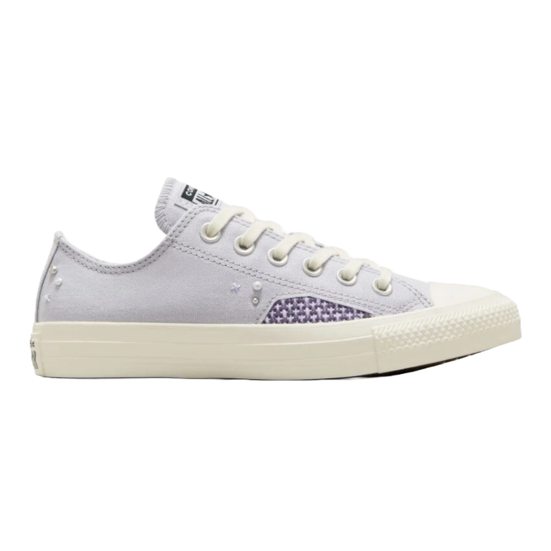 The Chuck Taylor Pearls & Studs Shoes are low-top sneakers featuring a light grey canvas upper with decorative pearl and stud embellishments. They have a textured rubber sole and a patch of purple checkered pattern on the side.