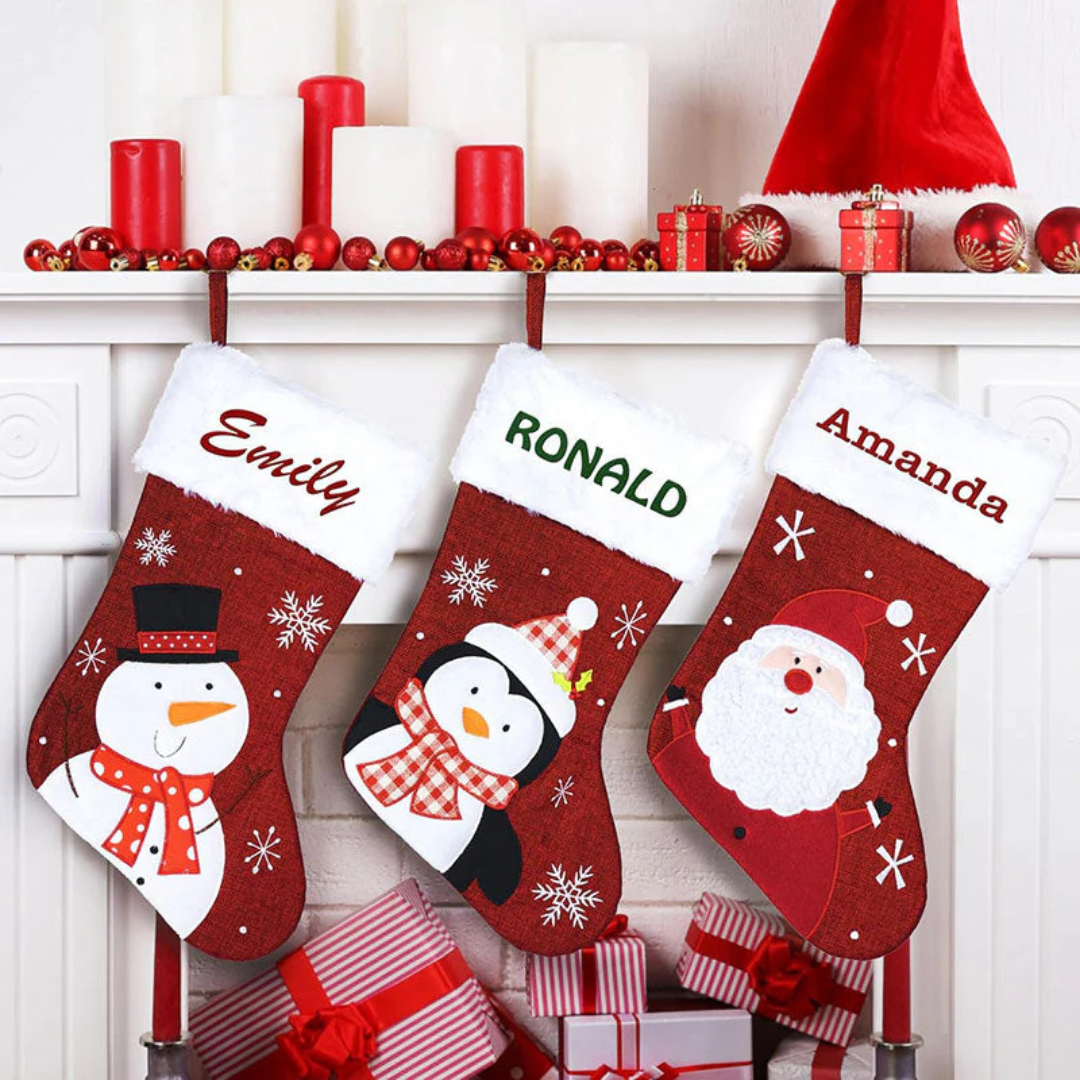Three Christmas stockings hanging on a mantel, each with a white top. They feature a snowman, a penguin, and Santa Claus design, labeled \“Emily,\“ \“Ronald,\“ and \“Amanda.\“ The mantel is decorated with red and white candles, ornaments, and small gift boxes.
