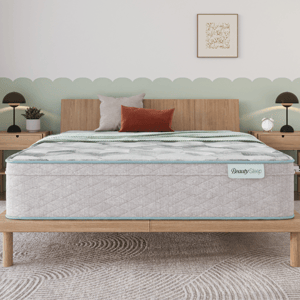A mattress labeled \“BeautySleep\“ on a wooden bed frame, accompanied by three decorative pillows, stands in a bedroom with light wood nightstands, modern lamps, and a patterned rug.