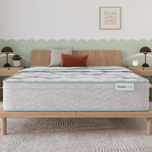 A mattress labeled \“BeautySleep\“ on a wooden bed frame, accompanied by three decorative pillows, stands in a bedroom with light wood nightstands, modern lamps, and a patterned rug.