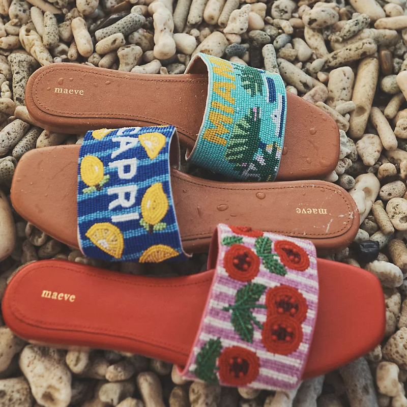 The Fiona Beaded Slide Sandals feature colorful embroidered straps with designs labeled \“CAPRI\“ with lemons, \“MIAMI\“ with palm motifs, and floral designs on pink stripes. They are by Maeve and rest on a rocky surface.