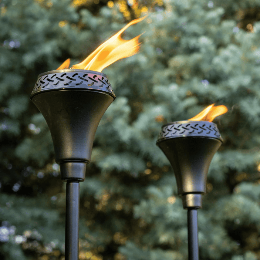 A pair of metal torches with a decorative perforated design are shown, with flames burning against a blurred background of greenery.