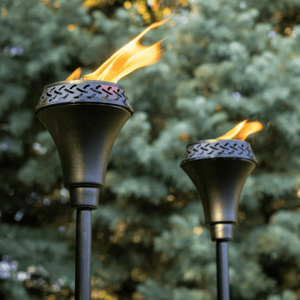A pair of metal torches with a decorative perforated design are shown, with flames burning against a blurred background of greenery.
