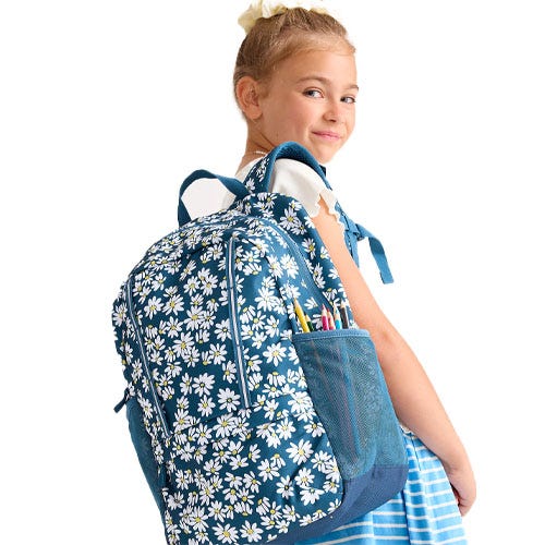 A young girl is wearing a blue backpack with a daisy pattern, carrying school supplies in its mesh side pocket. She is dressed in a blue and white striped skirt and a white top.
