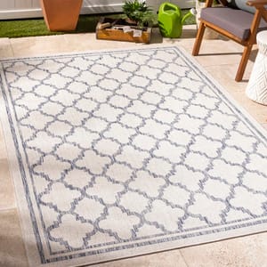 Outdoor rug with a geometric lattice pattern, laid on a patio with a chair, planter, tray, and watering can in the background.
