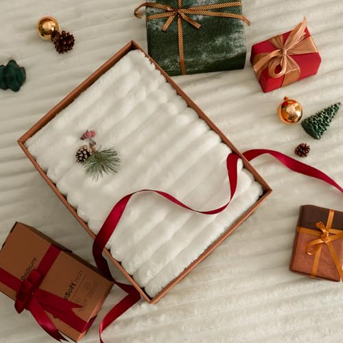 A gift box containing a white, soft fleece blanket with a red ribbon is surrounded by wrapped presents and small holiday decorations like pinecones and ornaments.