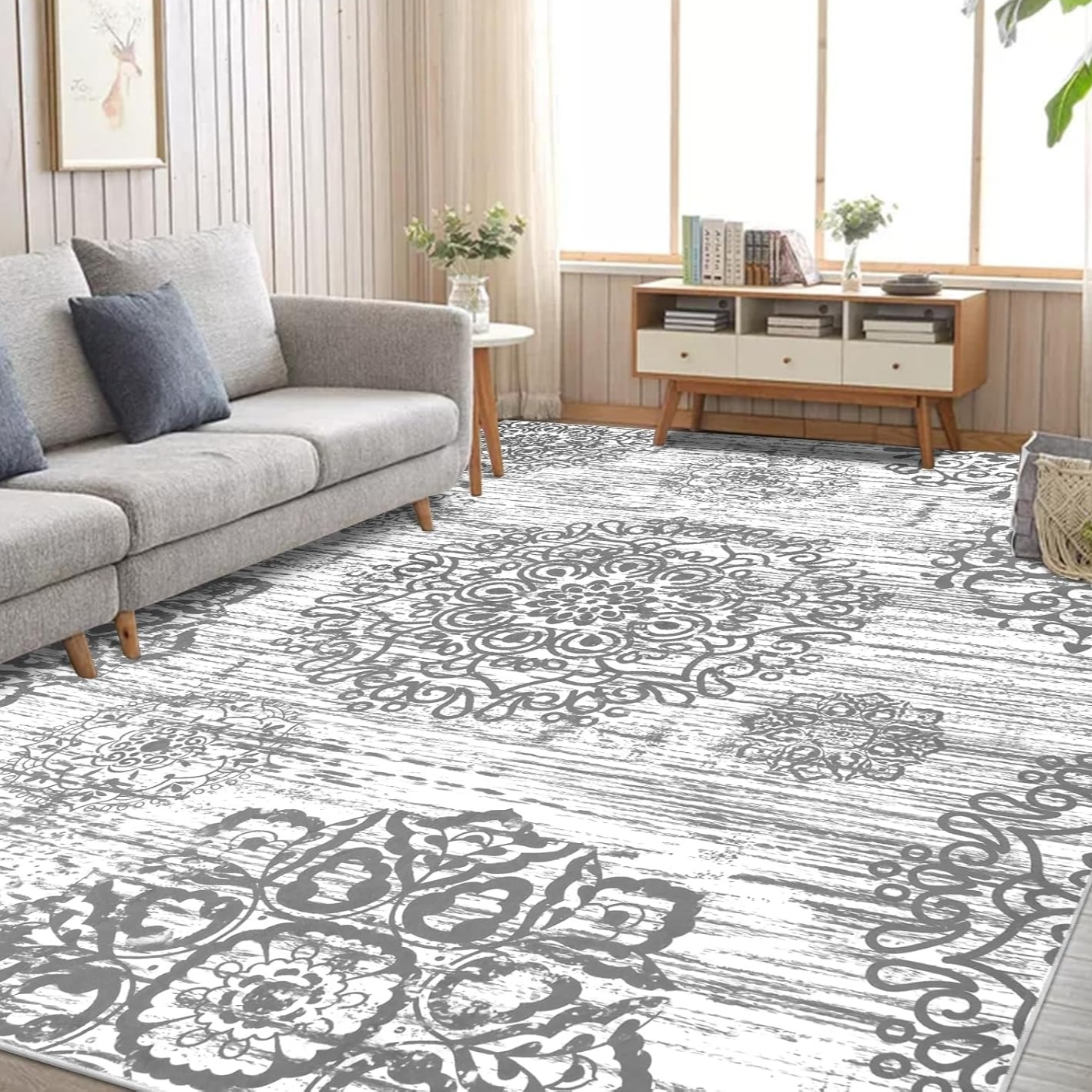 A living room features a gray sofa with blue cushions, a patterned gray and white area rug, a wooden console, and decorative items. Large windows with sheer curtains allow natural light to brighten the space.