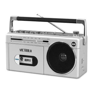 A silver Victrola boombox with a handle, cassette player, and speaker, featuring AM/FM radio and Bluetooth connectivity. The front panel displays frequency settings and a slot for cassette tapes.