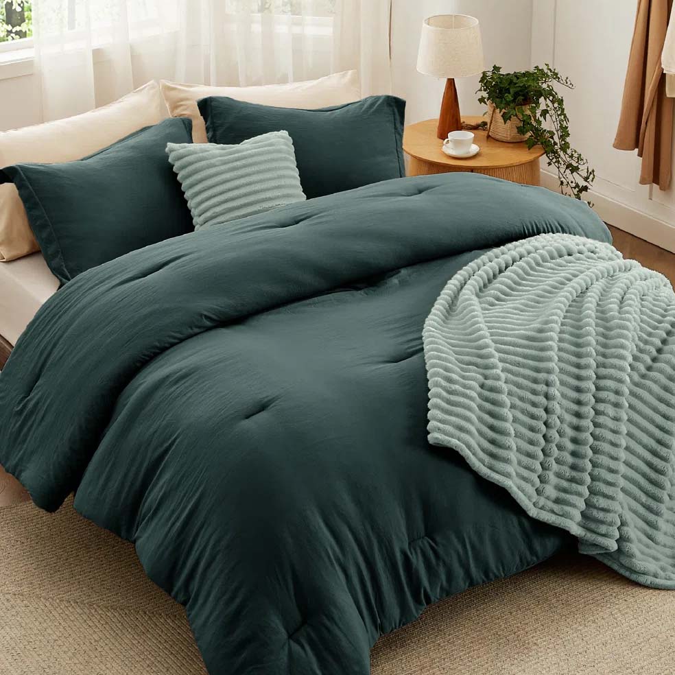A cozy bedroom setup featuring a dark green comforter, matching pillowcases, and a light green textured blanket. The scene includes a small bedside table with a lamp, plant, and cup.