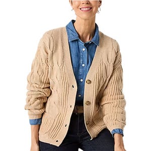 A woman wears a beige cable-knit cardigan over a denim button-up shirt, paired with dark jeans.