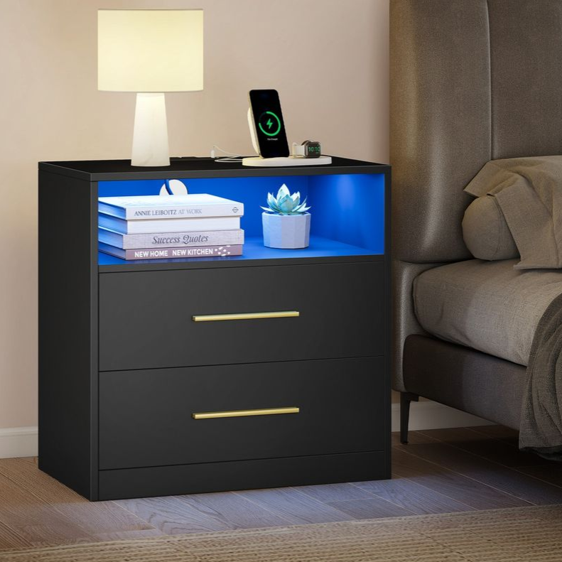 A black nightstand with a blue-lit open shelf, two drawers with gold handles, a lamp, and a charging phone and watch. Branded with \“VASAGLE by SONGMICS HOME.\“