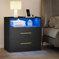 A black nightstand with a blue-lit open shelf, two drawers with gold handles, a lamp, and a charging phone and watch. Branded with \“VASAGLE by SONGMICS HOME.\“