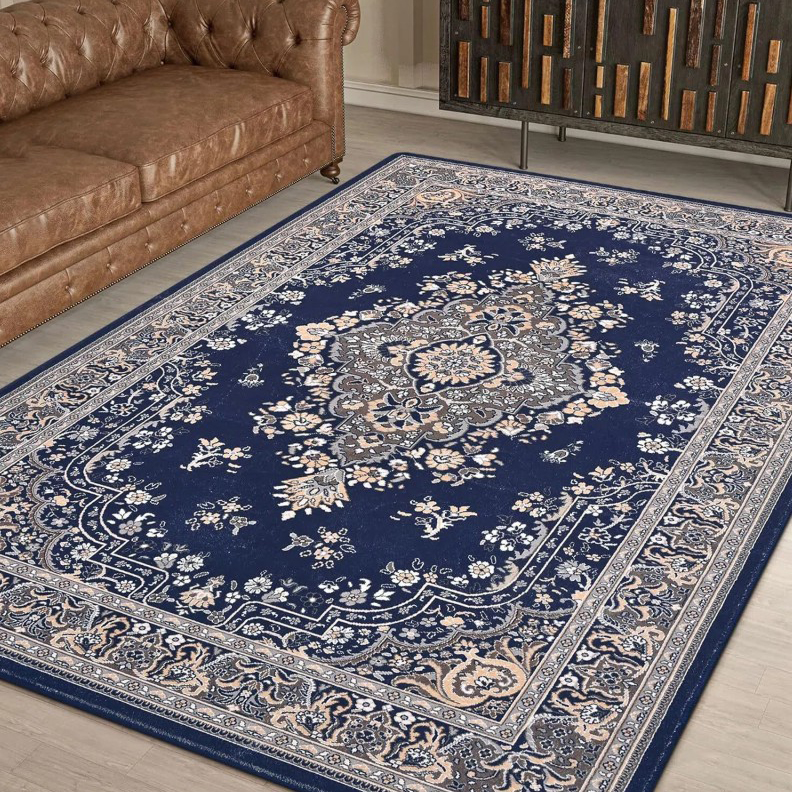 A blue Persian-style rug with intricate floral patterns, placed on a wooden floor, next to a brown tufted leather sofa.