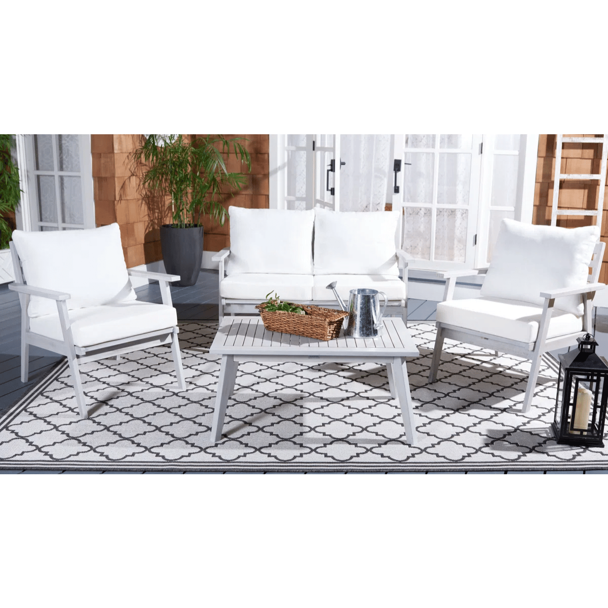 Outdoor patio set featuring two white cushioned armchairs, a loveseat, and a matching coffee table with a watering can and basket on top, placed on a patterned rug. There's a lantern in the foreground and potted plants in the background.