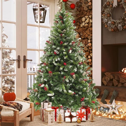 A decorated Christmas tree with red berries, pine cones, and wrapped gifts underneath stands in a cozy room with a fireplace, wood logs, and holiday wreaths.