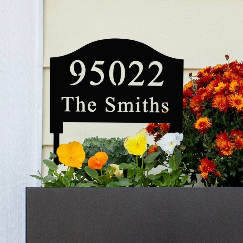 A black, personalized yard sign displays the number \“95022\“ and the name \“The Smiths,\“ positioned above a planter with colorful orange, yellow, and white flowers.