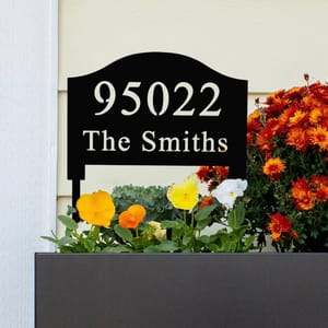 A black, personalized yard sign displays the number \“95022\“ and the name \“The Smiths,\“ positioned above a planter with colorful orange, yellow, and white flowers.