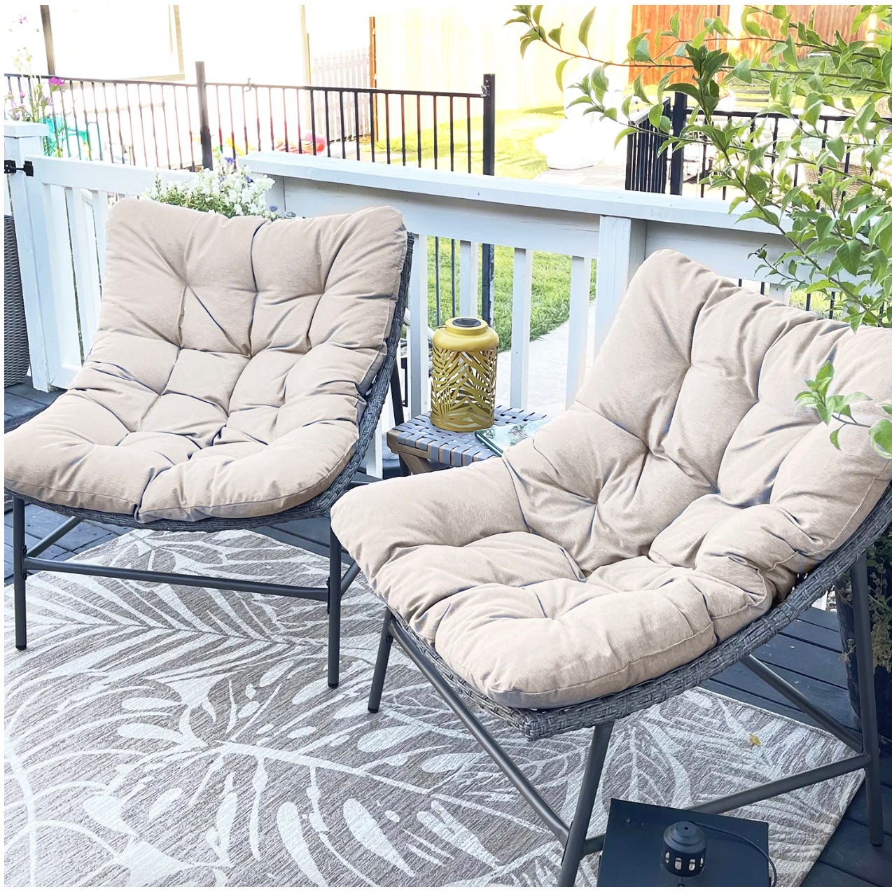Two tan cushioned patio lounge chairs set on a patterned outdoor rug, with a yellow decorative lantern on a small side table between them.