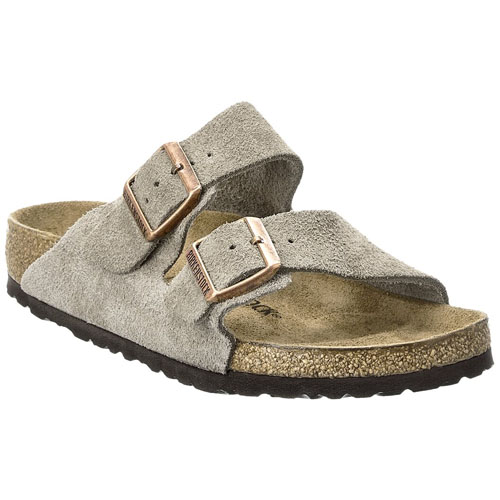 A beige suede sandal with two adjustable straps featuring brown buckles and a cork sole with a textured black bottom.