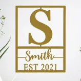Wall decor featuring a large letter \“S,\“ the name \“Smith,\“ and \“EST 2021,\“ with a minimalist design accompanied by plants on either side.