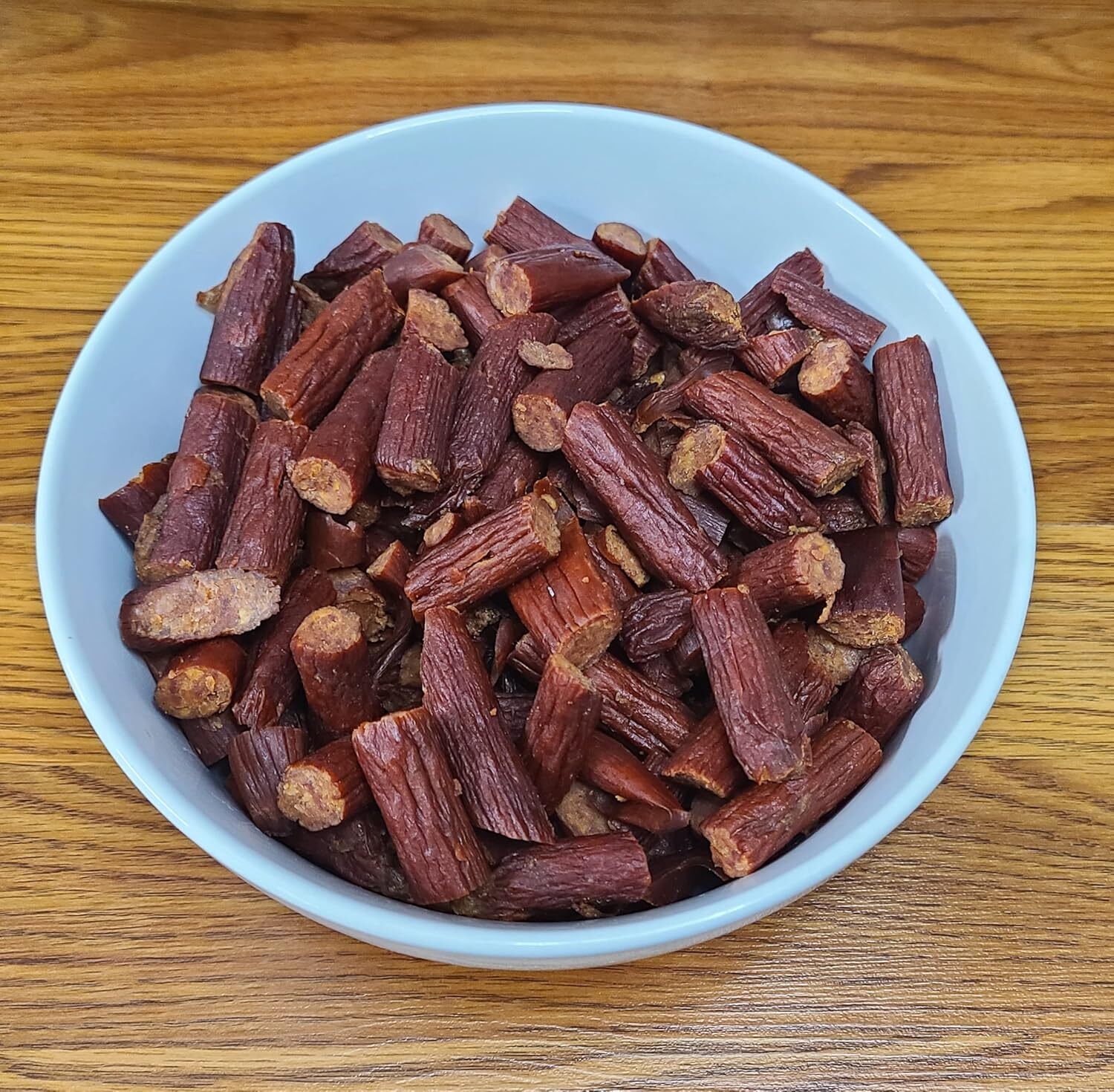 A bowl filled with small, dark brown pieces resembling chopped smoked sausages, placed on a wooden surface.