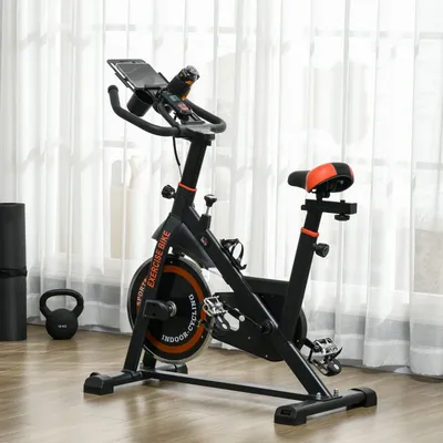 Indoor exercise bike with adjustable seat and handlebars, featuring a flywheel marked \“EXERCISE BIKE\“ and \“INDOOR CYCLING,\“ a tablet holder, and a water bottle holder. Nearby are a yoga mat and a kettlebell.