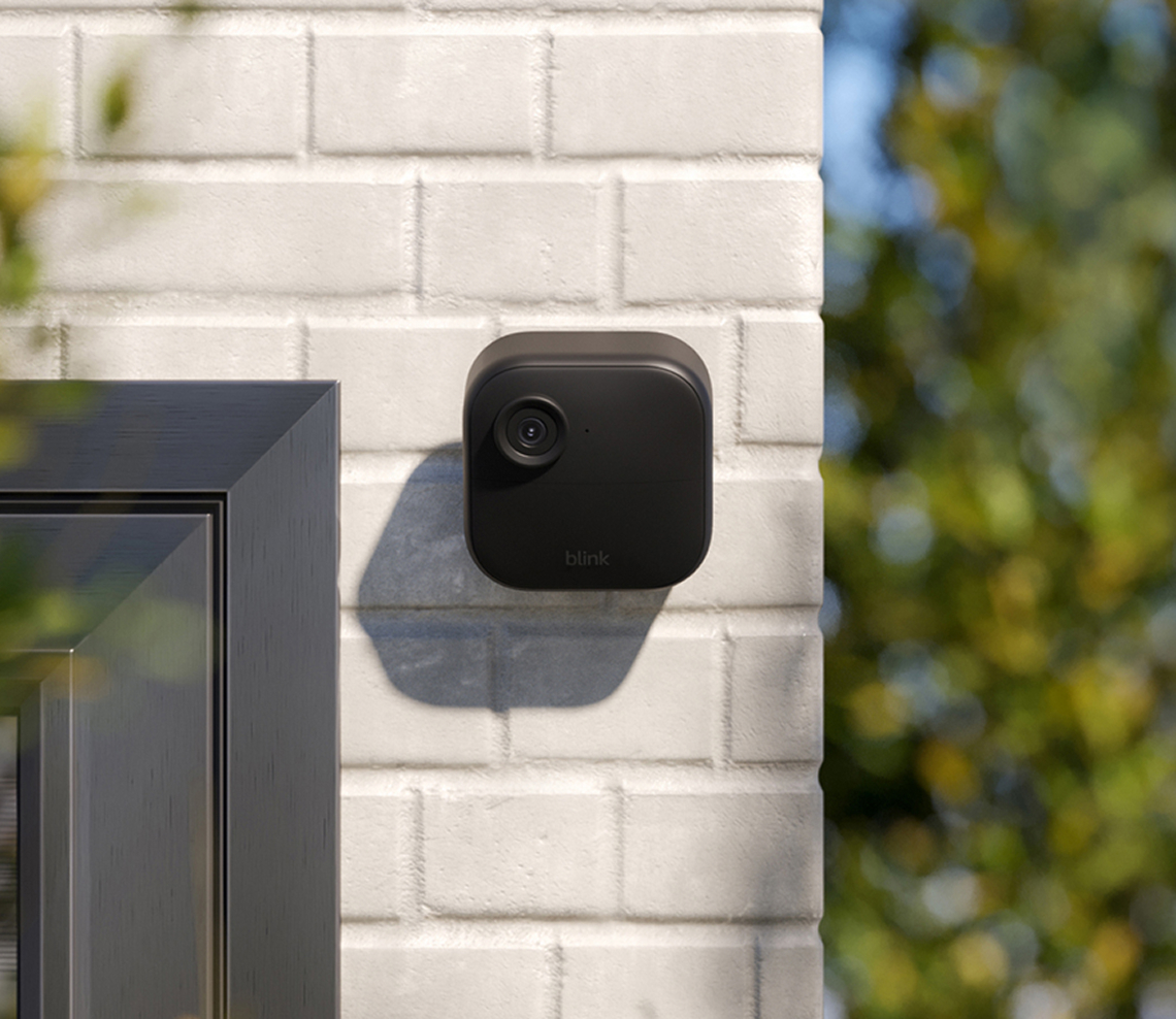A Blink outdoor security camera is mounted on a white brick wall near a window.