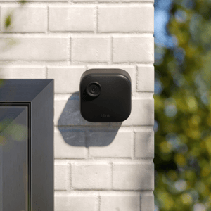 A Blink outdoor security camera is mounted on a white brick wall near a window.