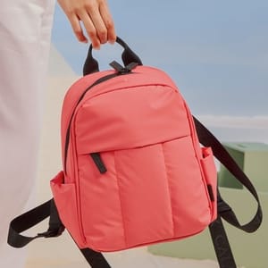 A small pink backpack with black straps and handles, shown against a beachy backdrop, held by a person in white clothing.