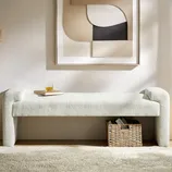 Modern upholstered bench with boucle fabric, topped with a book and drink, beneath abstract wall art. A woven basket containing books is placed underneath on a light carpeted floor.