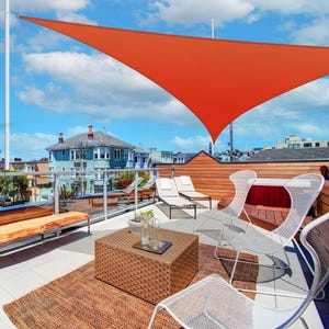 Rooftop patio with modern furniture: wicker-style coffee table, white mesh chairs, chaise lounges, and an orange triangular sun shade. Glass railing and a view of surrounding buildings.