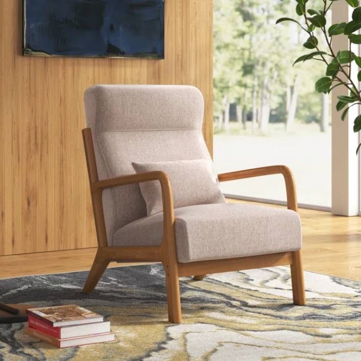 A mid-century modern armchair with wooden arms and beige upholstery is set on a patterned floor rug, with a stack of books nearby, in a room with wooden walls and large windows.