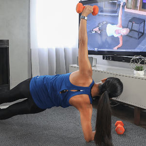 A person in a blue tank top exercises with orange dumbbells while following a workout video on a TV screen.