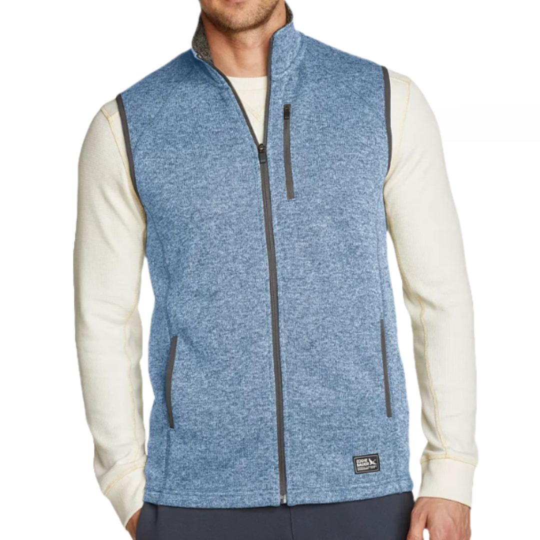 A man is wearing a blue zip-up vest over a long-sleeved cream-colored shirt. The vest features a high collar, zippered side pockets, and a chest pocket.