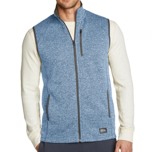 A man is wearing a blue zip-up vest over a long-sleeved cream-colored shirt. The vest features a high collar, zippered side pockets, and a chest pocket.