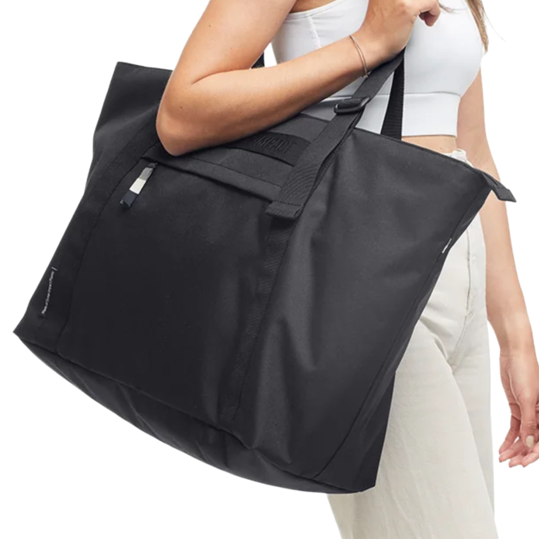 A person carries a large black tote bag with shoulder straps. The bag has a minimalist design with a small tag on the side.