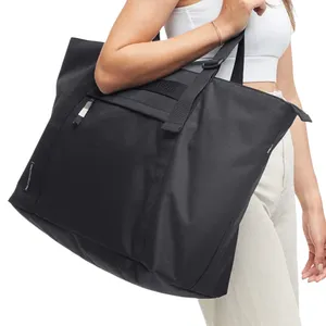 A person carries a large black tote bag with shoulder straps. The bag has a minimalist design with a small tag on the side.