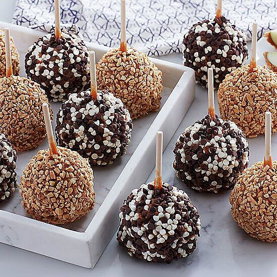 Assorted caramel apples coated with chopped nuts and chocolate chips, displayed on a white tray.