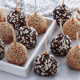 Assorted caramel apples coated with chopped nuts and chocolate chips, displayed on a white tray.