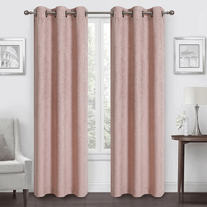 Room with pink curtains on a metal rod, flanked by two framed pictures. A lamp and flowers rest on a side table, with an armchair on the opposite side.