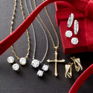 A collection of gold and silver jewelry featuring necklaces with pendants, including a cross, stud earrings with round stones, and hoop earrings, displayed with a red velvet ribbon and gift box.