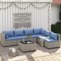 Outdoor wicker sectional sofa set with blue cushions, including a corner piece and a square glass-top coffee table. Background includes a wall with string lights and macramé art.
