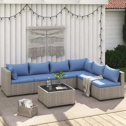 Outdoor wicker sectional sofa set with blue cushions, including a corner piece and a square glass-top coffee table. Background includes a wall with string lights and macramé art.