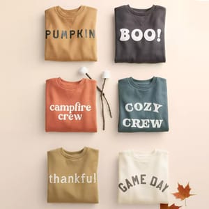 Six folded sweatshirts with different colors and text; phrases like \“PUMPKIN,\“ \“BOO!,\“ \“campfire crew,\“ \“COZY CREW,\“ \“thankful,\“ and \“GAME DAY\“ are featured.