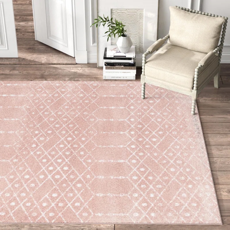Soft pink 5' x 7' area rug with a white geometric diamond pattern, placed on wooden flooring next to a light upholstered chair and a stack of books with a white vase.