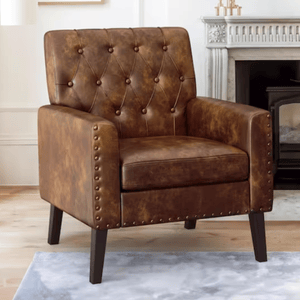 A brown leather armchair with a tufted backrest and nailhead trim, featuring wooden legs, set in a living room with a fireplace in the background.