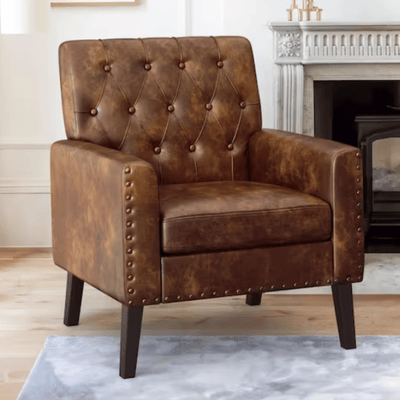 A brown leather armchair with a tufted backrest and nailhead trim, featuring wooden legs, set in a living room with a fireplace in the background.