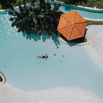 A person relaxes on a float in a large, clear pool next to green plants and a small structure with an orange-tiled roof.