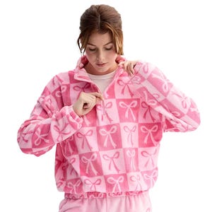 A pink plaid quarter-zip pullover featuring a soft, plush texture and a bow pattern. The garment includes elastic cuffs and hem, offering a cozy and snug fit.
