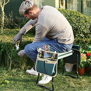 A man kneels on a dual-purpose gardening aid that can serve as a seat when flipped over, equipped with side pockets for holding tools.